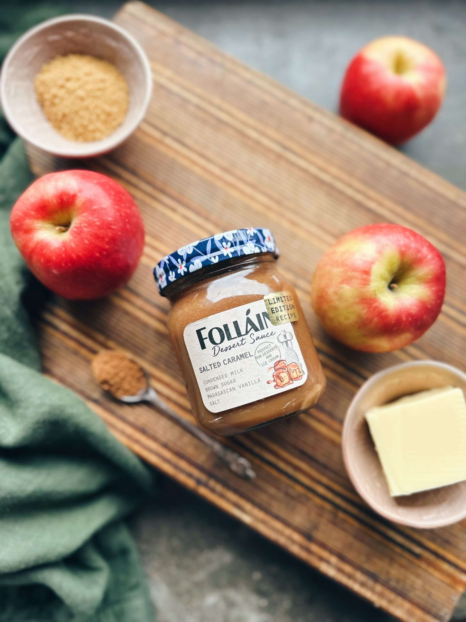 jar of follain irish cream liqueur lying on side on wooden chopping board surrounded by red apples bowl of butter and bowl of brown sugar